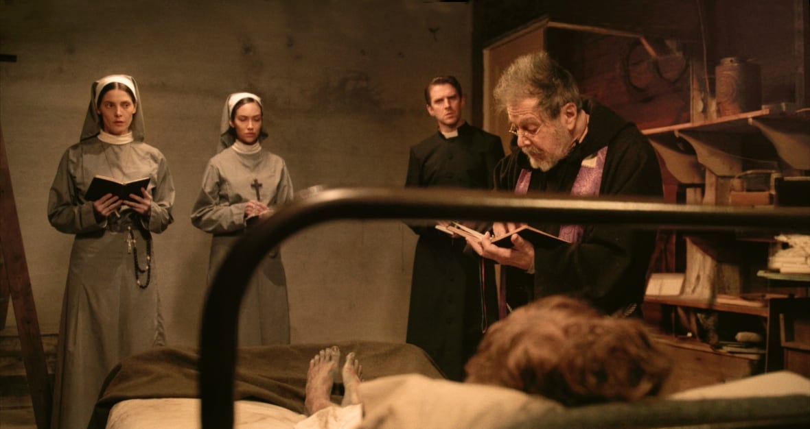 A priest reading from a book stands in front of a woman in a bed and beside two nuns and another priest.. Only the woman's feet are visible, which are bare and dirty.