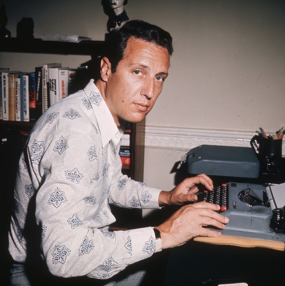 A dark haired, cleanshaven man sits near a typewriter in a photo that appears several years old.