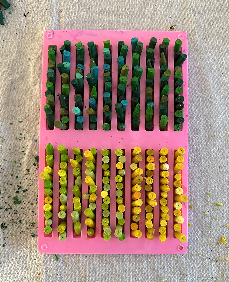 A silicone mold with broken pieces of crayons in green and yellow color families, ready for the oven.
