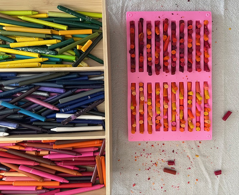 A divided container of old crayons on a table with a silicone mold with broken crayons bits, ready to make new crayons in the oven.