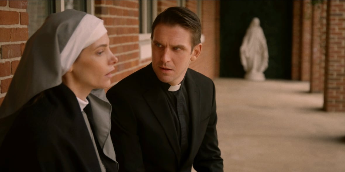 A priest speaks to a nun. They are both seated in an outdoor hallway.