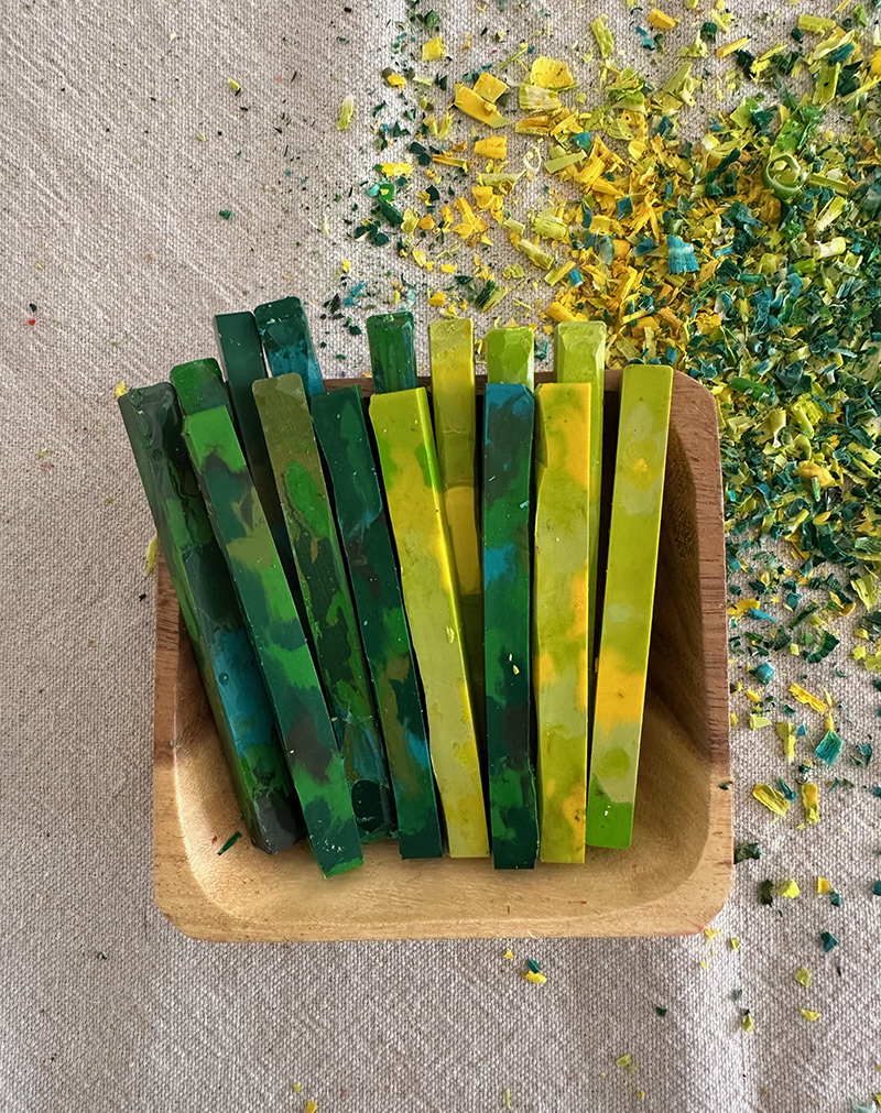 A small wooden bowl holding a set of homemade crayons in green and yellow hues.
