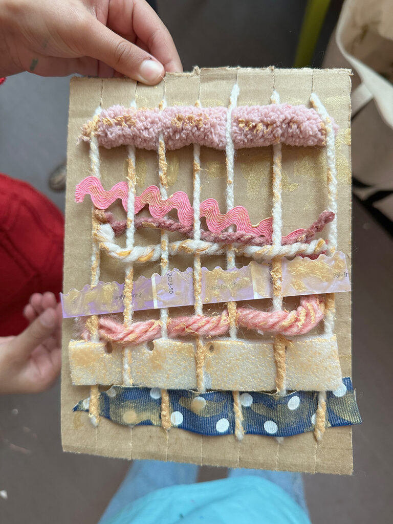 Child holding up their finished cardboard loom weaving that they have embellished with gold paint.