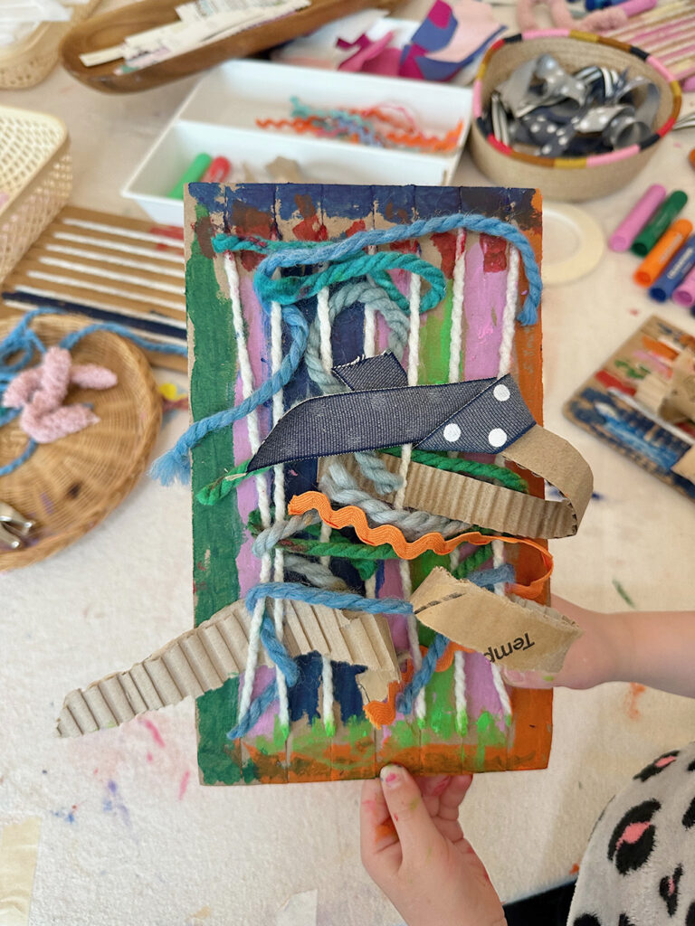 Child holding up their cardboard loom weaving where they have used lots of different materials and textures, like yarn and ribbon and strips of corrugated cardboard.